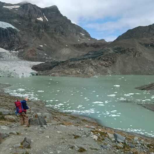 Un lago in alta montagna, con un ghiacciaio sullo sfondo. In primo piano, un ricercatore con uno zaino capiente che contiene il robot per le analisi